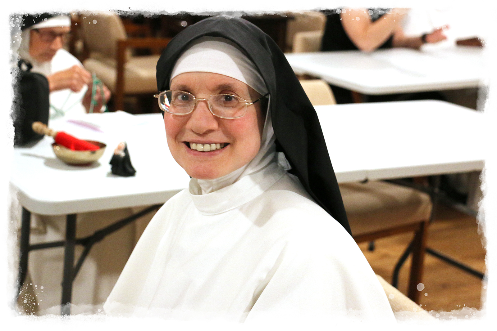 A smiling nun wearing a traditional black and white Dominican habit with glasses, seated at a white folding table at the Monastery of Mary the Queen. Other people and tables are visible in the blurred background, suggesting a community gathering or event